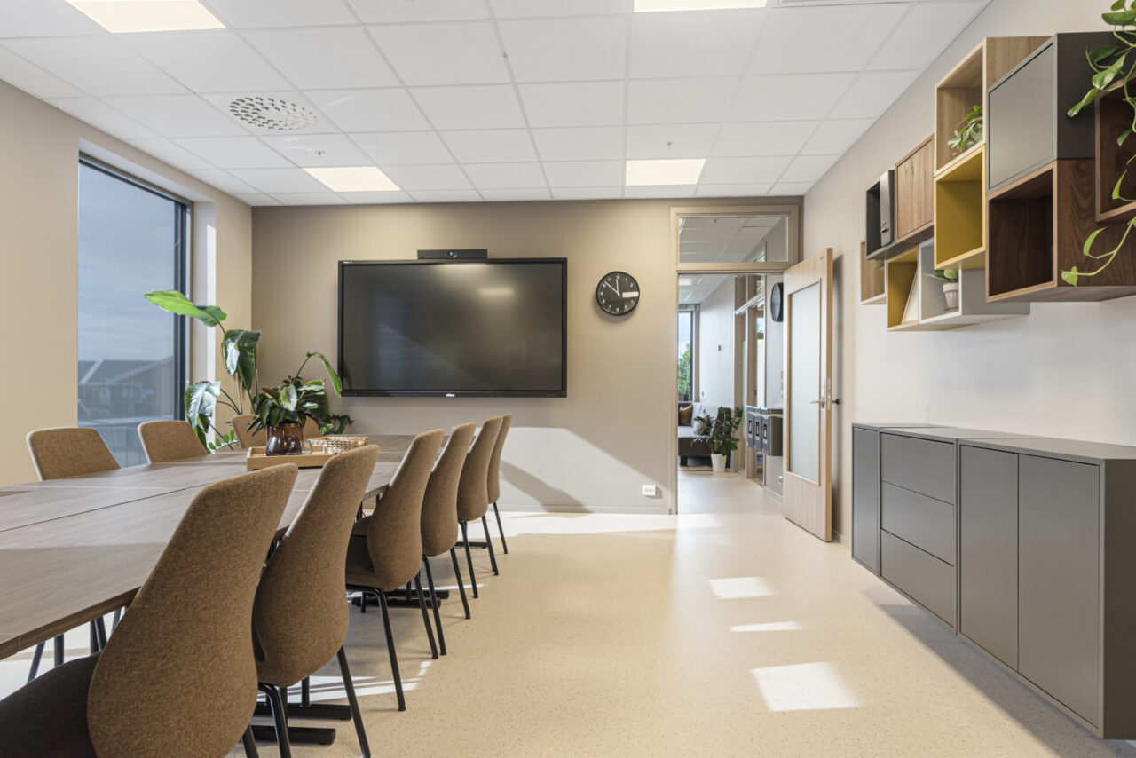 Møterom med bord og stoler, tv på veggen og hyller med blomster på den ene veggen