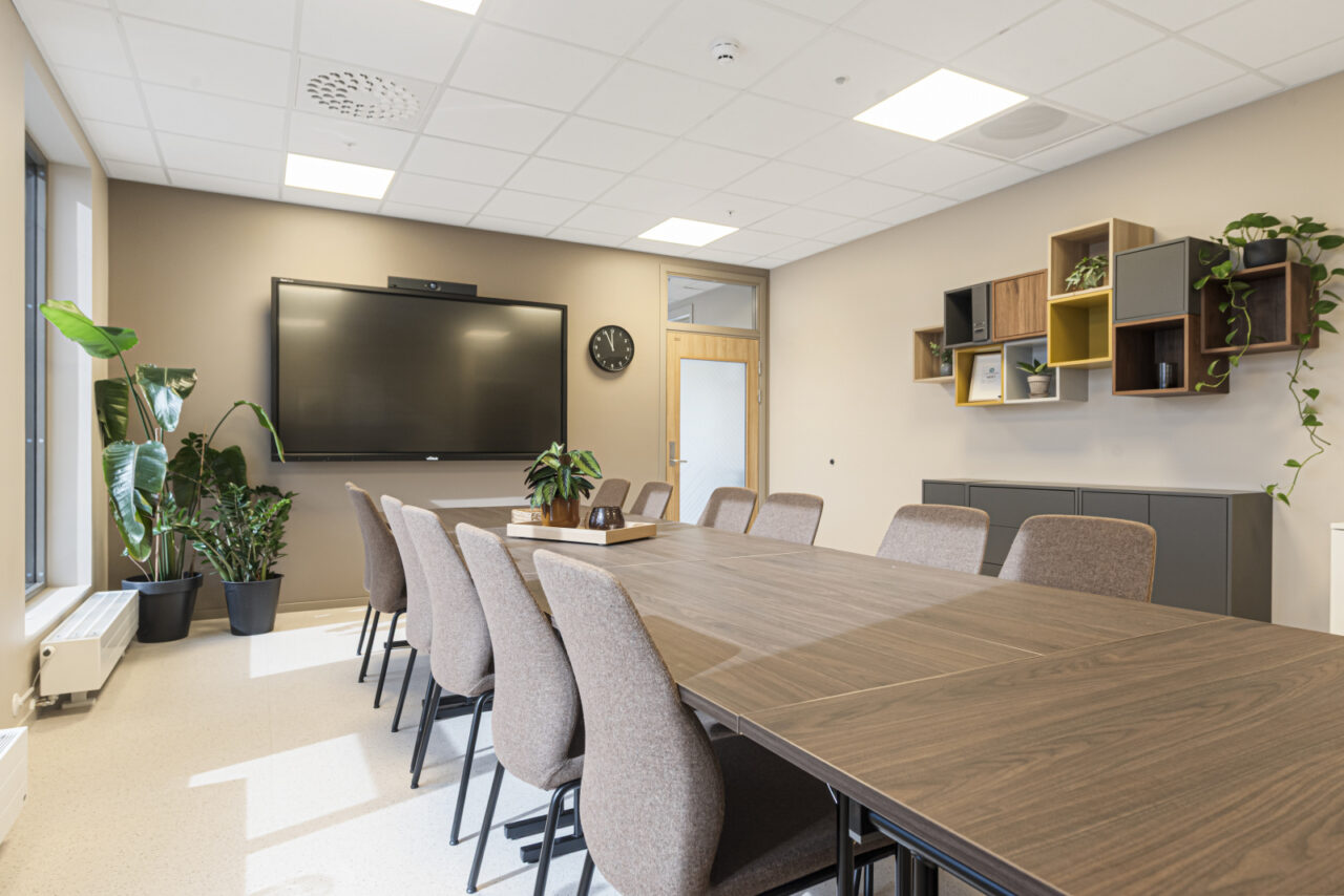 Møterom med bord og stoler, tv på veggen og hyller med blomster på den ene veggen