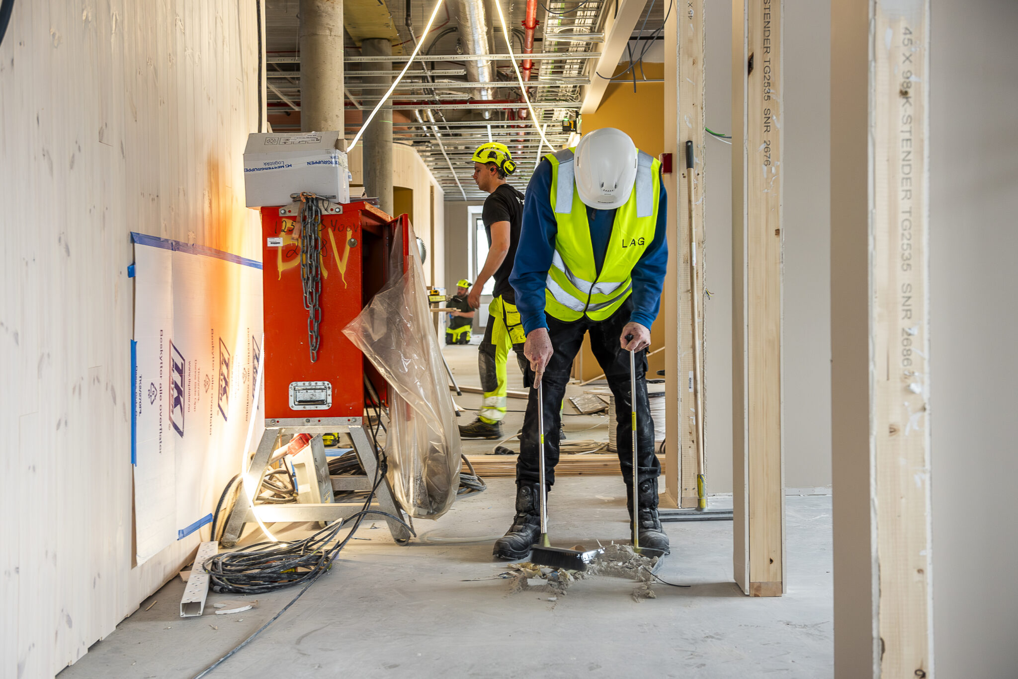 mann med kost og støvbrett på en byggeplass. i bakgrunnen ser du to andre tømrere.