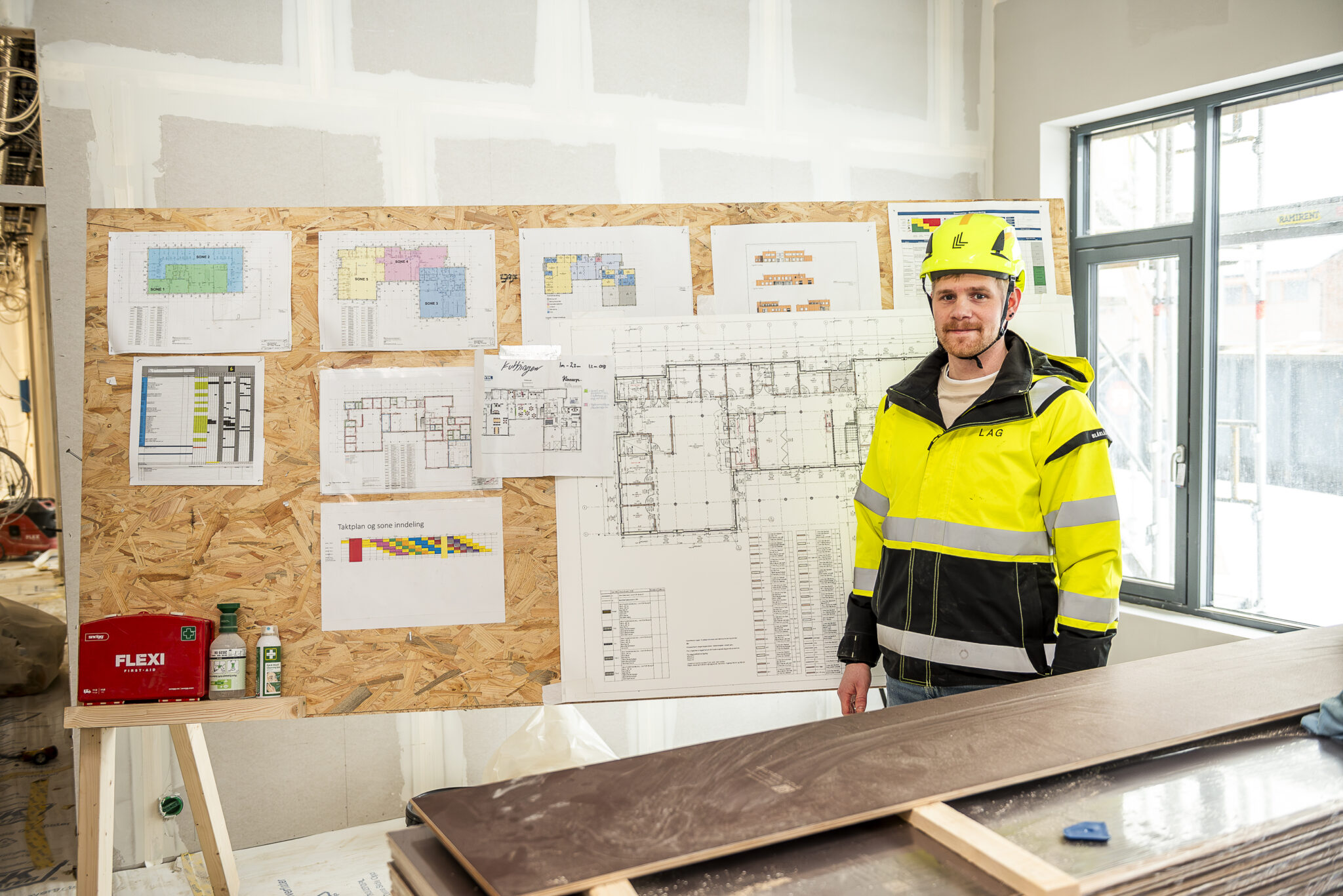 Smilende mann i arbeidsklær foran en tavle med arkitekttegninger og planer