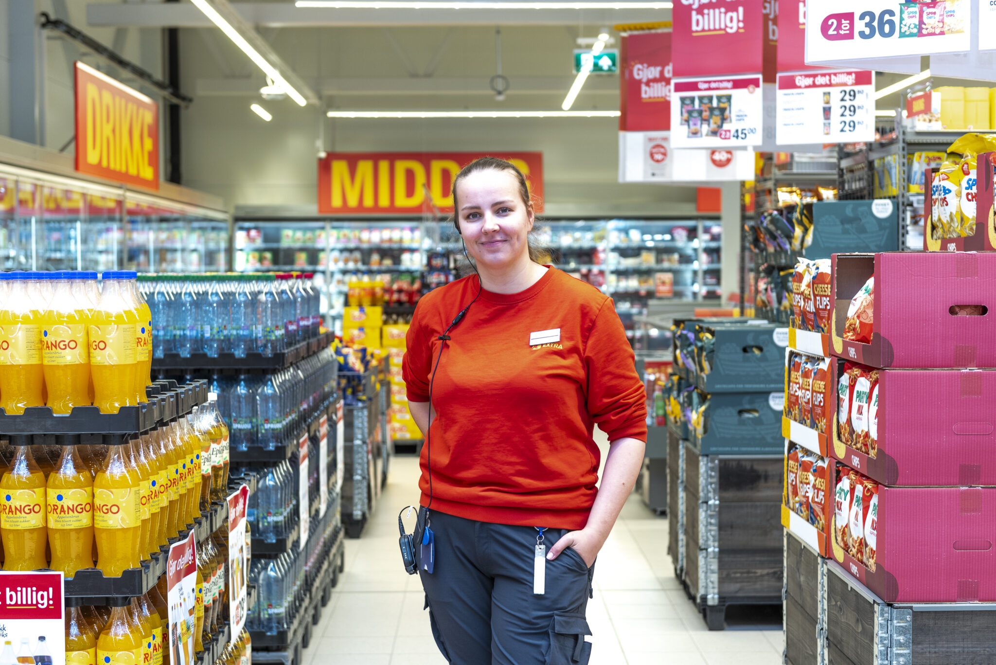 Smilende dame i arbeidsklærne til Coop Extra står mellom butikkhyller