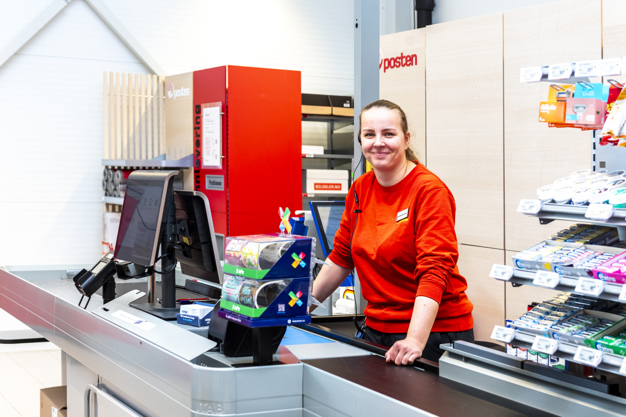 Smilende dame i arbeidsklær står bak kassa i en butikk