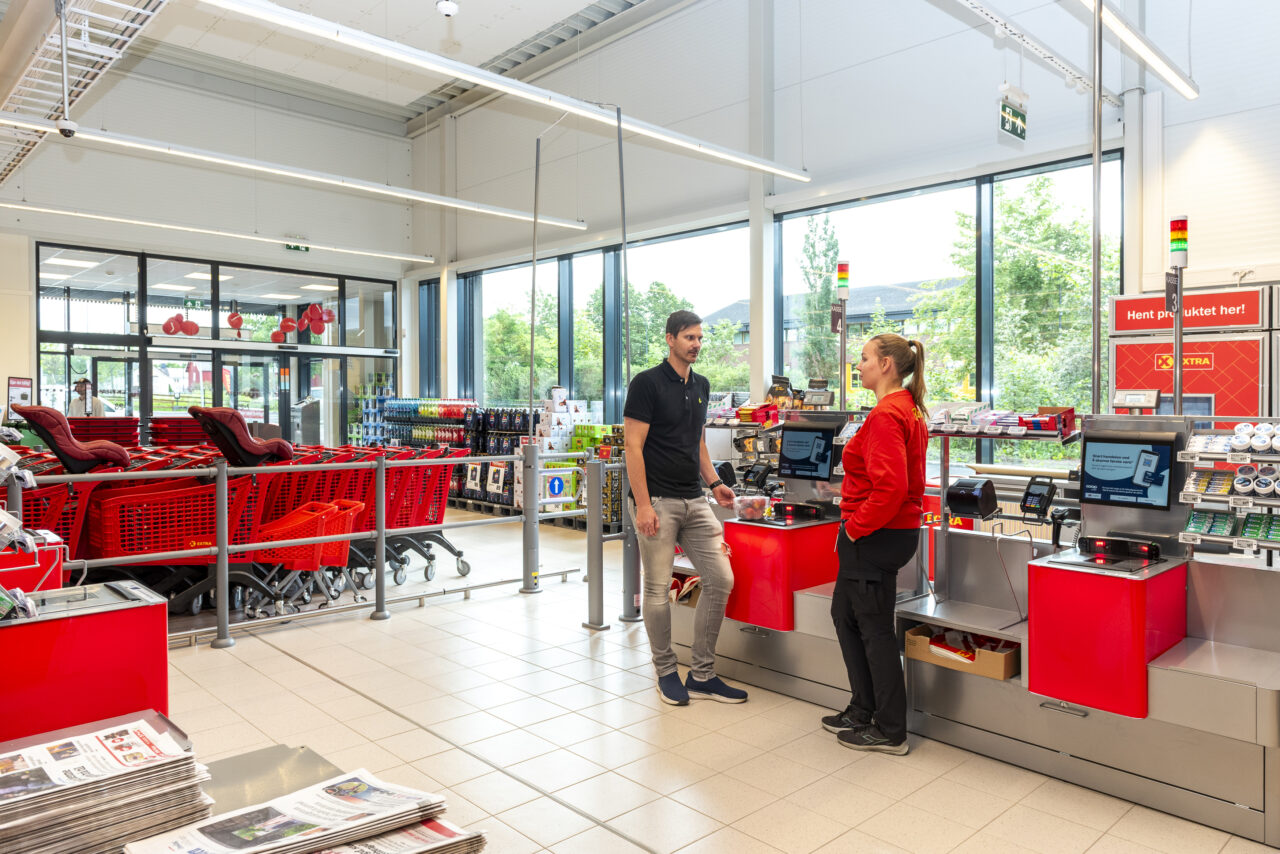 En mann og en dame står og snakker sammen foran selvbetjeningskassen i en matbutikk. bak dem er det store vinduer.