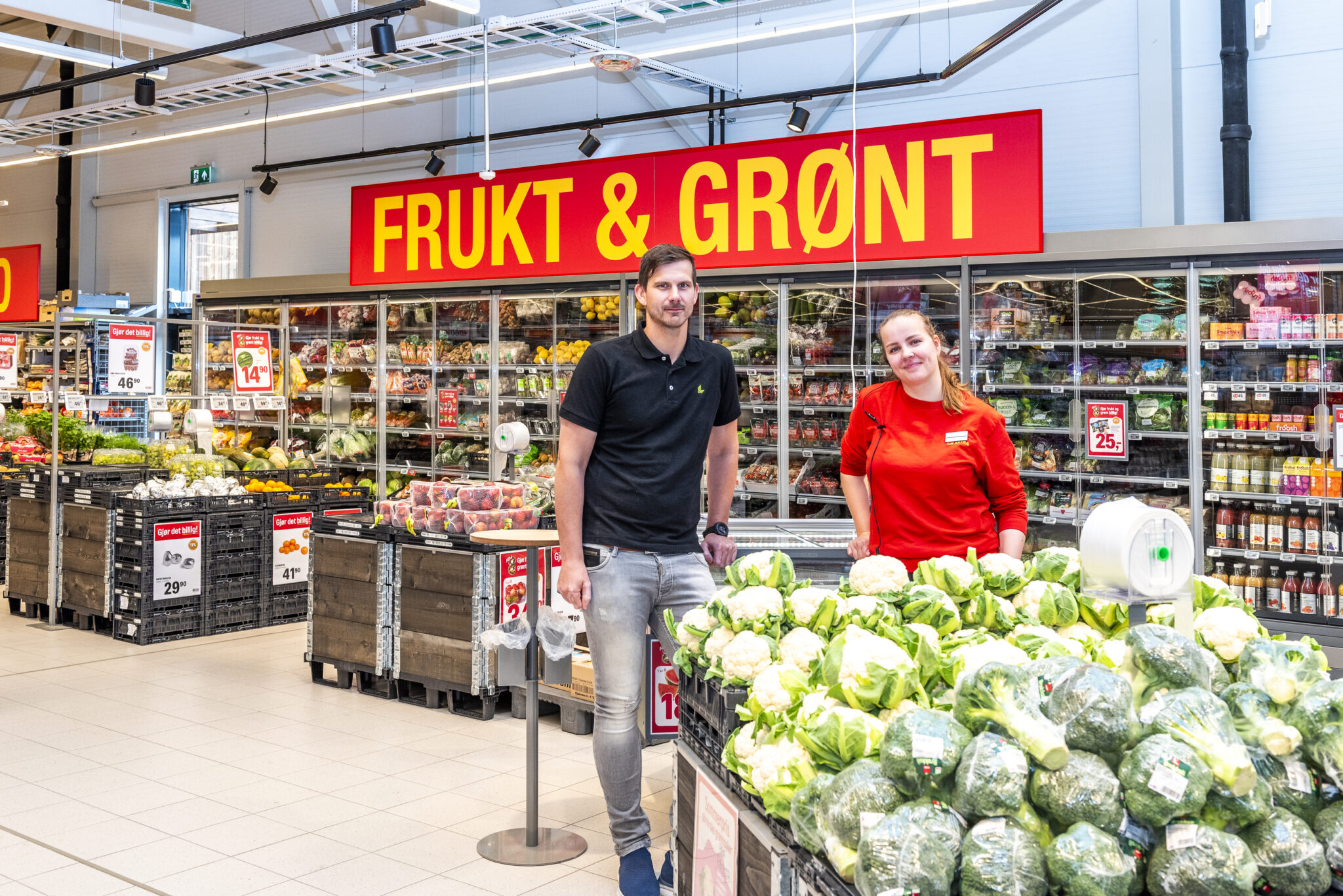 En mann og en dame stor foran et skilt det står Frukt & grønt på og bak en hylle med blomkål.